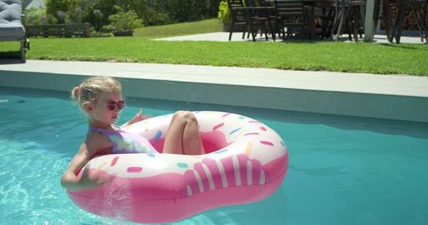 Girl lounging on pink donut float in backyard pool wearing red sunglasses