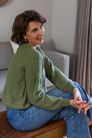 Relaxed woman sitting on ottoman wearing green knit sweater and jeans in cozy living room