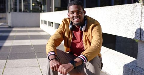 Smiling African American man sitting on plaza ledge wearing headphones and mustard jacket
