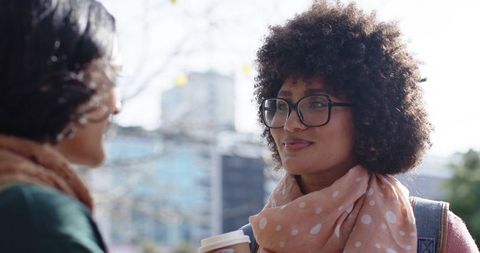 Smiling multicultural women talking on city street while holding coffee and wearing scarves