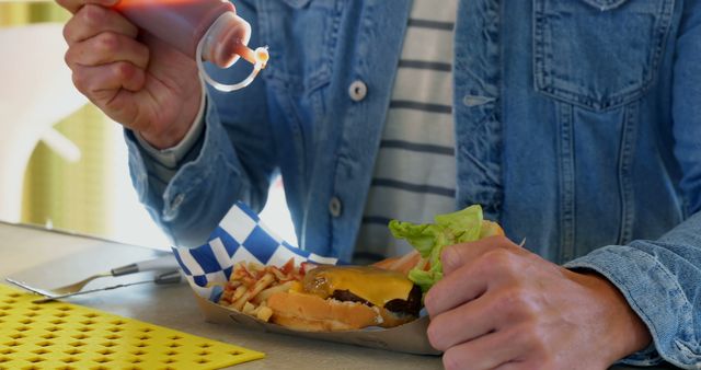 Person in a casual setting adding sauce to a cheeseburger while holding it. Tray with fries visible. Ideal for use in advertisements for fast food, casual dining, food delivery services, kitchenware related to food preparation, or health and wellness articles discussing diets or eating habits.