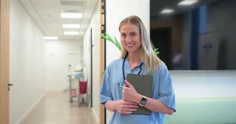 Smiling female clinician holding tablet and stethoscope in modern clinic corridor