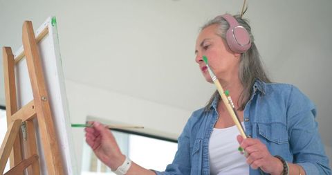 Mature woman painting on canvas at easel using dual brushes wearing headphones and denim shirt