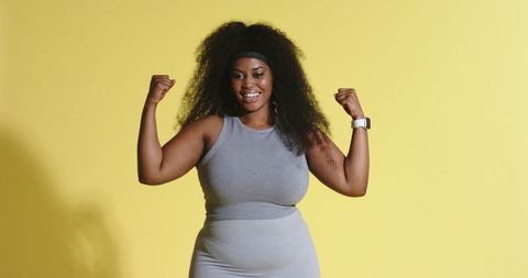 African american woman flexing muscles smiling wearing smartwatch on yellow backdrop