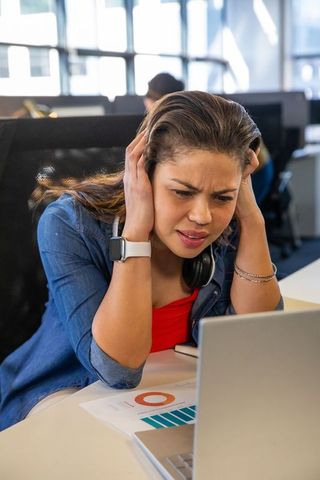 Stressed woman covering ears while reviewing noisy report on laptop in open plan office