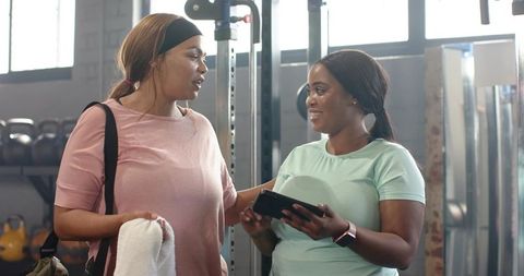 Two diverse women chatting in gym holding tablet and towel, workout motivation in weight area
