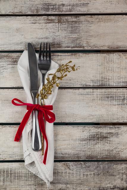 Elegant cutlery wrapped in a white napkin tied with a red ribbon and decorated with golden sprigs, placed on a rustic wooden plank. This setting is perfect for fine dining, holiday parties, festive meals, and casual dining aesthetics. It can be used in advertisements, food blogs, restaurant menus, and event planning.