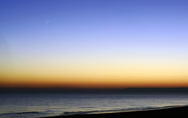 Image shows a crescent moon hanging in a gradient twilight sky transitioning from blue to orange, reflecting on the calm sea, perfect for backgrounds, meditation visuals, or nature-themed projects.