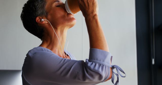A mature woman savoring a hot cup of coffee while listening to music with earphones. She appears relaxed and indulgent in her casual attire during a break. Ideal for lifestyle articles, blog posts about relaxation, mindfulness, or the benefits of taking small breaks during work or activities.