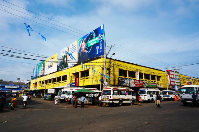 This image shows a lively marketplace with a prominent yellow building covered in various advertisements. The street is busy with white vans, buses, and people going about their day. Ideal for use in tourism promotions, urban studies, articles about bustling city life, or illustrating commercial activity.
