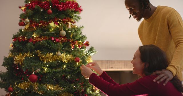 Happy diverse couple decorating christmas tree at home, slow motion. Relationship, togetherness, christmas, tradition, celebration, domestic life and lifestyle, unaltered.