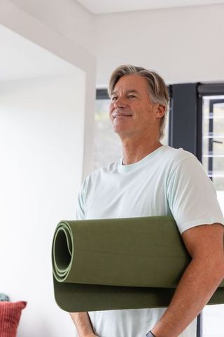 Mature man holding green yoga mat standing by window, home yoga wellness lifestyle portrait