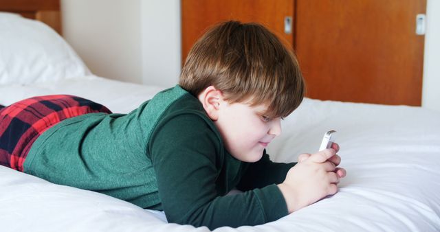 Young boy in casual clothing lying on a bed while playing with a smartphone. Scene depicts a comfortable indoor setting, ideal for usage in themes related to childhood, technology, leisure, and modern lifestyle. Suitable for articles, advertisements, and blogs discussing children's use of technology, safe use of mobile devices, or home environments.