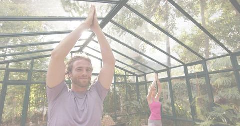 Couple practicing yoga together in sunlit glass greenhouse surrounded by tropical plants