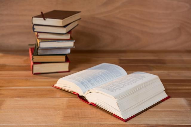 Open book on wooden table with stack of books in background. Ideal for educational content, library promotions, study guides, academic articles, and literature-themed projects.