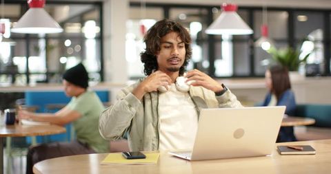 Young man working on laptop in modern coworking lounge with headphones and smartphone