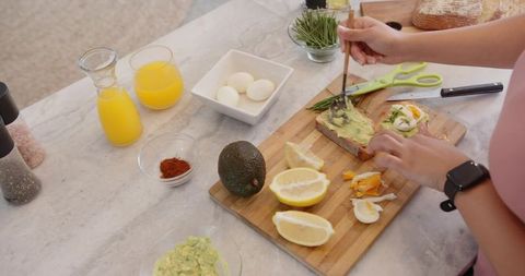 Adult woman spreading avocado on toast with boiled eggs lemon and herbs on cutting board