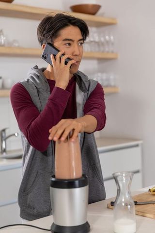 Young asian man making smoothie while talking on smartphone in modern kitchen