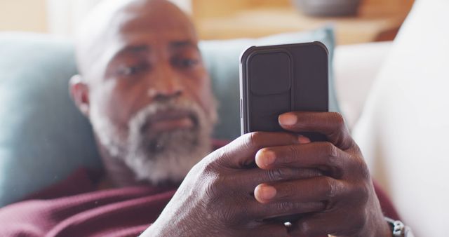 Elderly man sitting on a sofa, attentively using a smartphone. Highlighting modern technology use among senior adults. Suitable for topics on aging, technology adoption by older generations, and relaxed home environments. Can be used for blogs, articles, and campaigns focused on senior lifestyle and tech savviness of elderly people.