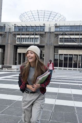 Laughing female student carrying notebooks and backpack while walking across campus plaza