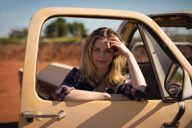 Young woman leaning on an open car door in a rural setting, enjoying a sunny day. Ideal for use in lifestyle blogs, travel articles, advertisements for outdoor activities, or automotive promotions.