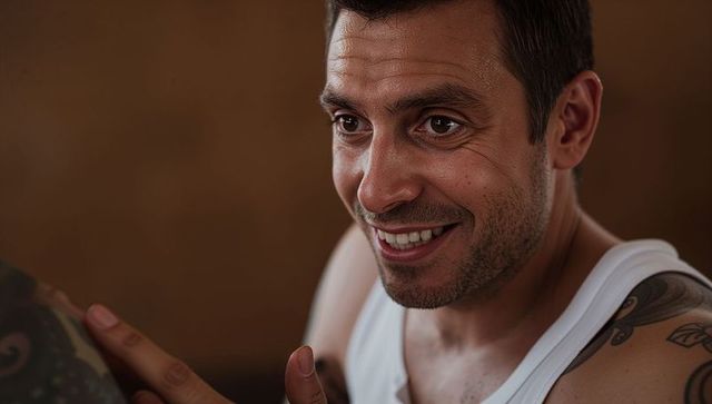 Smiling man wearing white tank showing arm tattoo and stubble warm closeup portrait