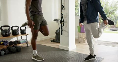 Diverse male friends stretching and balancing on one leg in home gym with kettlebells