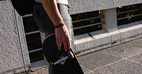 Skateboarder standing holding board by concrete railing on sunlit urban walkway
