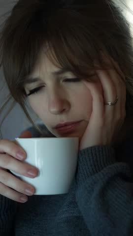 Contemplating woman cradling mug in gray sweater, blinking — vertical close-up video