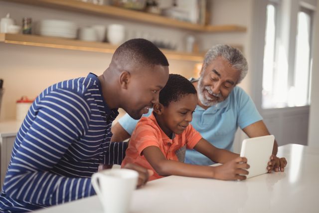Three generations of a family are gathered in the kitchen, engaging with a digital tablet. The grandfather, father, and son are smiling and enjoying their time together. This image can be used to depict family bonding, the use of technology in education, and the joy of spending time with loved ones. Suitable for advertisements, family-oriented content, and educational materials.