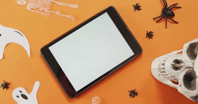 Tablet with blank screen amid Halloween decorations like skull, spiders, skeletons, and ghosts on orange background. Perfect for tech advertisements, Halloween promotions, app mockups, seasonal greetings.