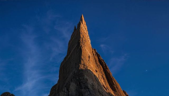 Sunlit granite spire piercing blue alpine sky with crescent moon and wispy cirrus clouds