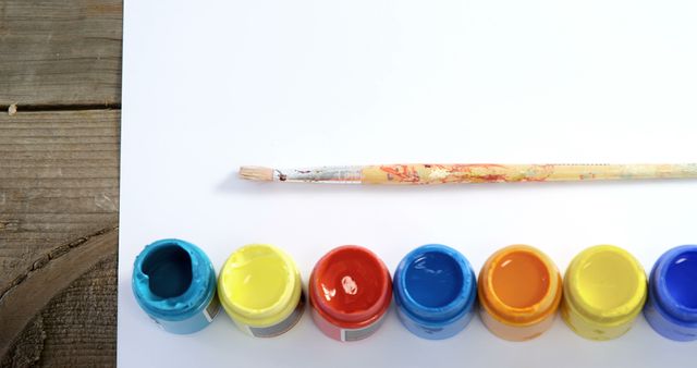 A paintbrush rests above a row of colorful paint pots on a wooden surface, with copy space. Art supplies like these inspire creativity and are essential for artists and hobbyists to express their imagination.