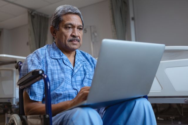 This image depicts a mature male patient in a wheelchair using a laptop in a hospital ward. It can be used for healthcare and medical-related content, patient care, technology in healthcare, elderly care, and rehabilitation services. It is suitable for articles, blogs, and promotional materials focusing on patient recovery, hospital facilities, and the integration of technology in healthcare.
