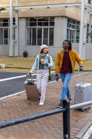 Young travelers walking with rolling suitcases holding coffee and phone in city plaza