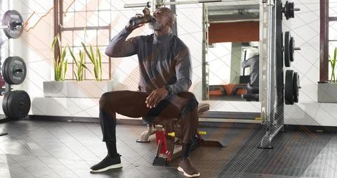 Mature athlete drinking water while resting on bench in modern gym with squat rack
