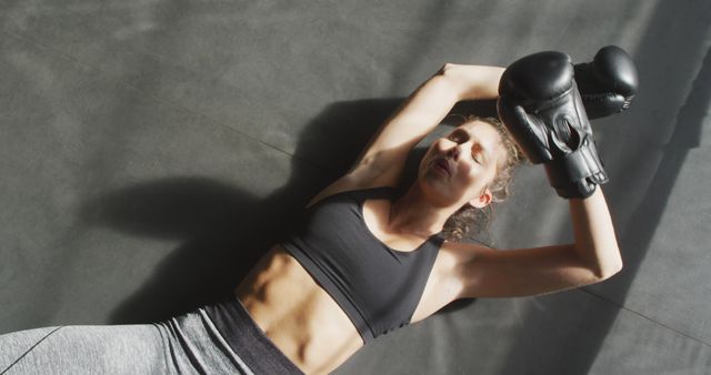 Female boxer is lying on the floor after intense workout, displaying exhaustion and effort. Useful for fitness blogs, sports articles, promotional materials for gyms and boxing academies, motivational content advocating for perseverance and dedication in sports.