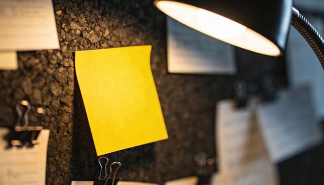 Yellow sticky note glowing on corkboard under warm desk lamp light for reminders