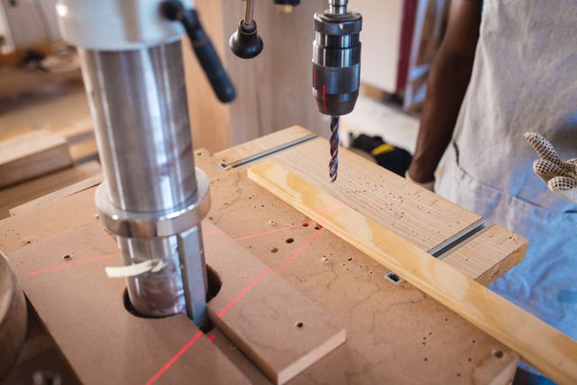 Carpenter operating a drill press in a workshop, focusing on precision drilling. Ideal for illustrating woodworking, carpentry skills, craftsmanship, and industrial manufacturing processes. Useful for articles, blogs, and advertisements related to DIY projects, professional carpentry, and woodworking tools.