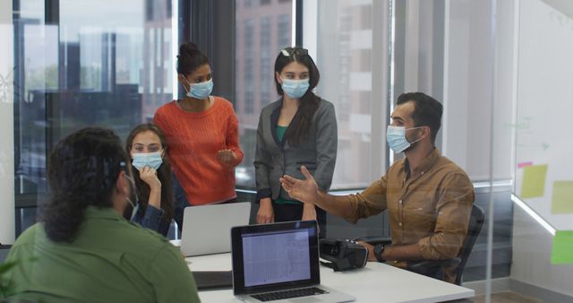 Group of coworkers collaborating in a modern office, discussing ideas and using laptops while wearing face masks. This depicts the new normal in the workplace during a pandemic, highlighting safety measures and teamwork. Ideal for articles on office safety, workplace collaboration, modern office environments, and pandemic-related topics.