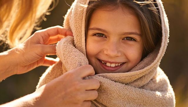 Smiling child wrapped in beige towel with helping hands, sunlit backyard family moment