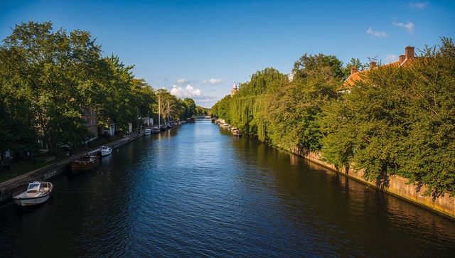 Sunlit canal framed by weeping willows and moored boats leading to distant spire