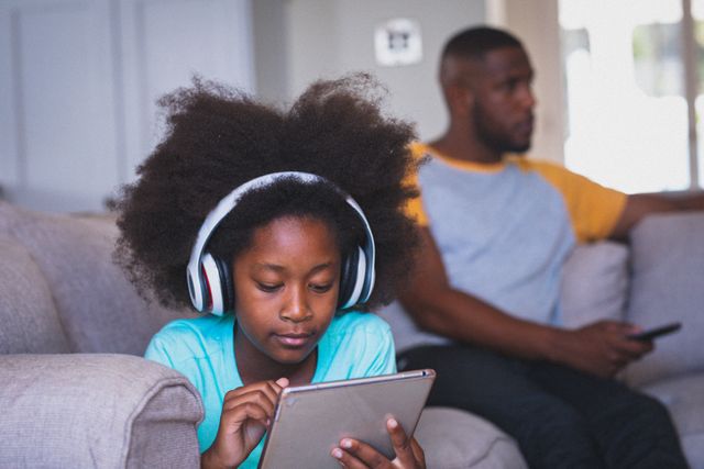 This image depicts an African American girl wearing headphones and using a tablet while her father watches TV in the background. It is ideal for illustrating themes related to family bonding, modern technology use, quarantine life, and parent-child relationships. Suitable for articles, blogs, and advertisements focusing on family dynamics, digital learning, and home entertainment.