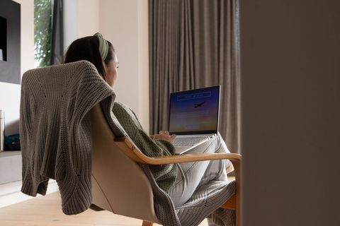 Asian woman booking flight on laptop in cozy minimalist living room wearing green sweater