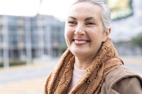 Smiling mature woman wearing knit brown scarf and tan coat on urban sidewalk, autumn portrait