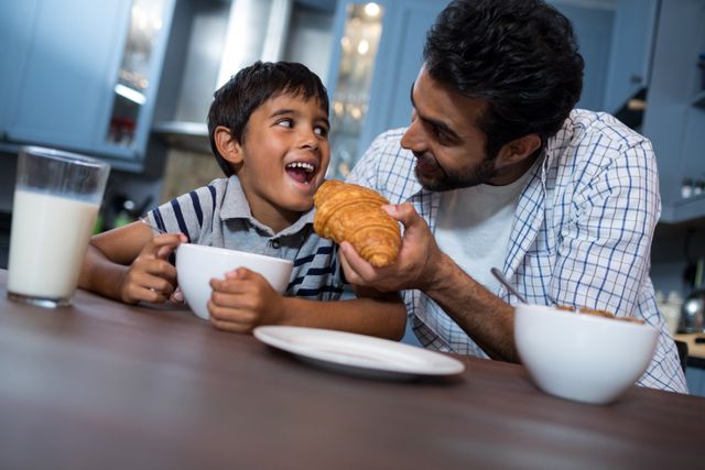 Smiling father feeding croissant to son while having breakfast
