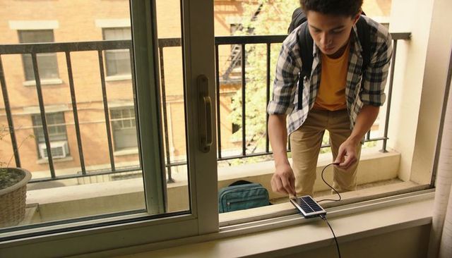 Teen connecting portable solar charger on apartment balcony windowsill with backpack