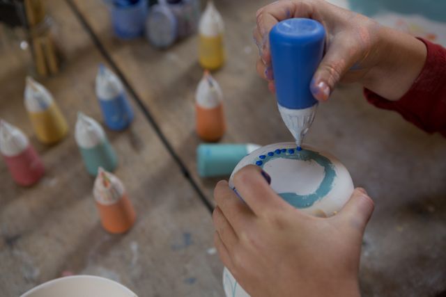 Boy decorating mug with paint in pottery workshop. Hands holding paint bottle, adding blue dots to ceramic mug. Various colorful paint bottles on table. Ideal for content related to children's activities, creativity, art and craft tutorials, DIY projects, pottery classes, and artistic hobbies.
