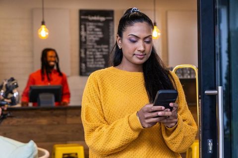 Asian woman checking smartphone while entering cozy cafe in mustard sweater, barista in background