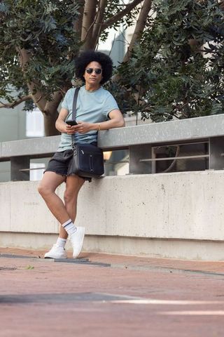 African man leaning on railing using smartphone wearing crossbody bag casual streetwear